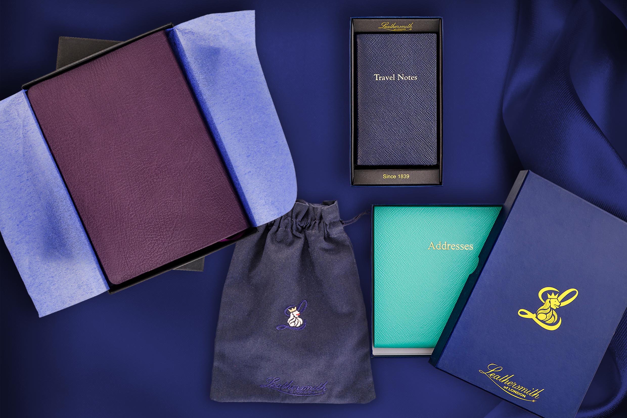 Flat lay of Leathersmith stationery gift set arranged on deep navy fabric. An open presentation box reveals a burgundy leather notebook wrapped in tissue paper