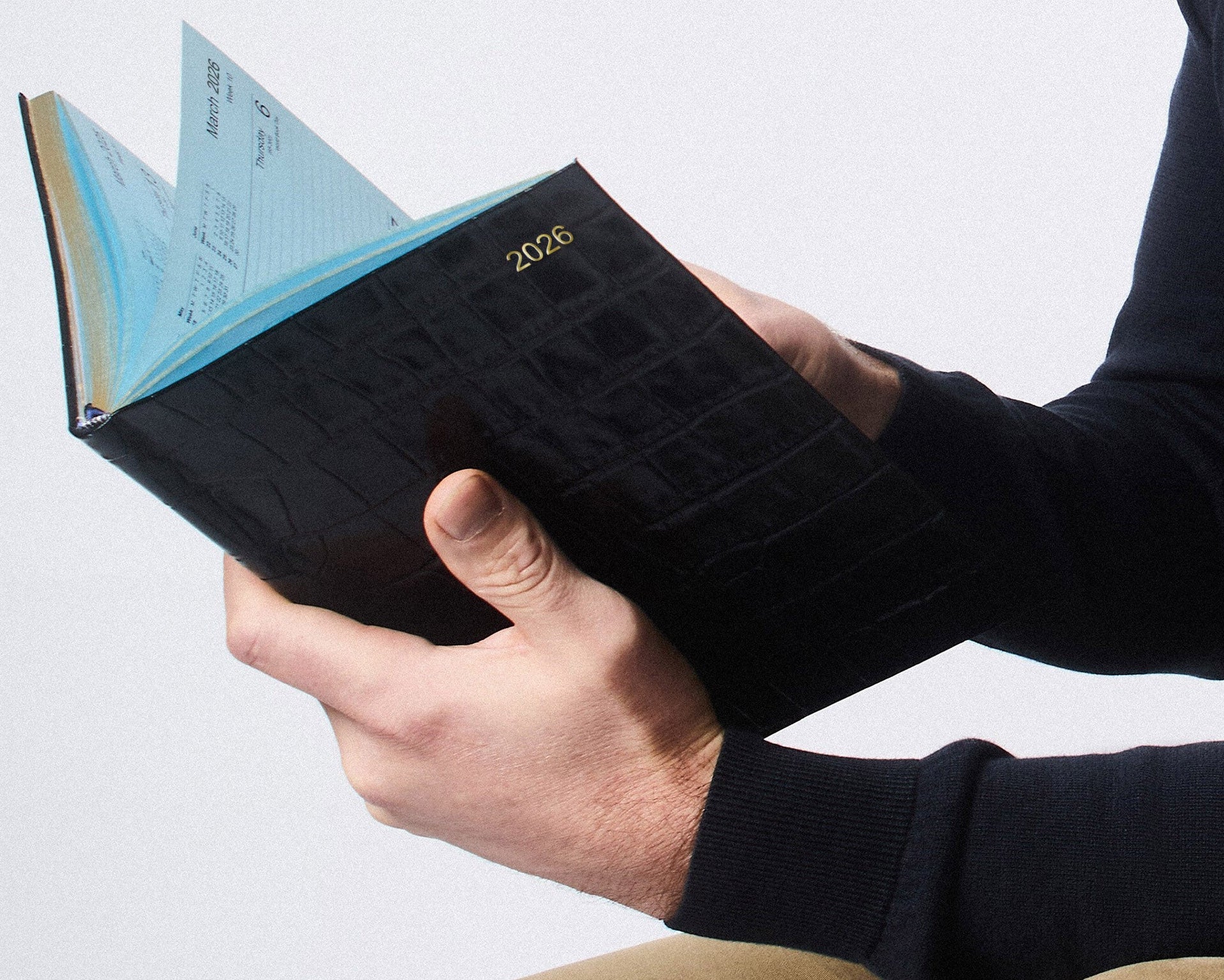 Hands holding an open 2026 Leathersmith diary with a black croc-textured leather cover, gold embossed year detail, and signature light blue pages.